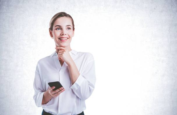 Portrait of smiling young businesswoman using smartphone near concrete wall. Concept of communication. Mock up - Foto, Bild
