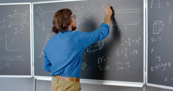 Rear of Caucasian mannelijke docent schrijven wiskunde of natuurkunde formules met krijt op schoolbord. Achteraanzicht van een leraar die op school werkt. Les wiskundekunde. Portret van een glimlachende wiskundige. - Video