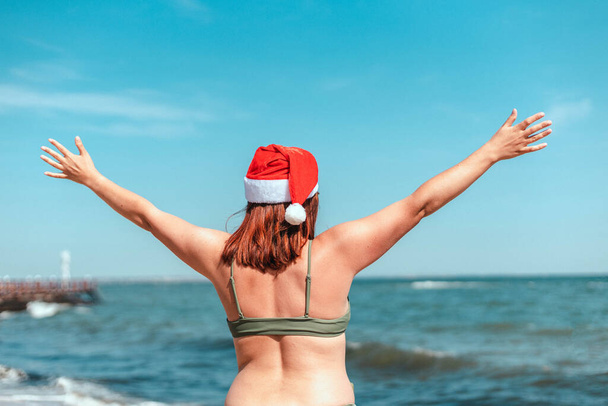Vacaciones de Navidad en un país cálido. Chica joven con la espalda en un sombrero de santa y un traje de baño con las manos extendidas mira al mar. - Foto, Imagen
