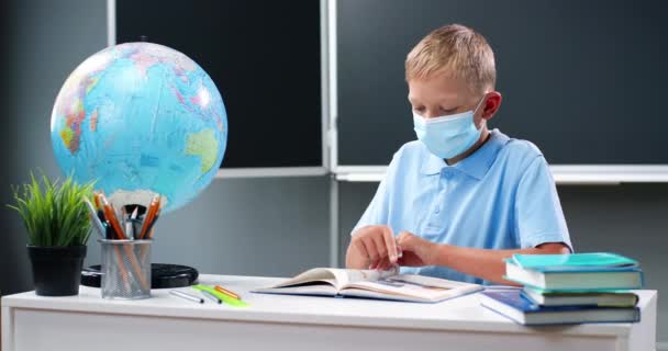 Small Caucasian teen boy in medical mask sitting at desk with world globe in school. Teenage schoolboy reading textbook and flipping pages. Pupil learning in classroom. Coronavirus concept. - Footage, Video