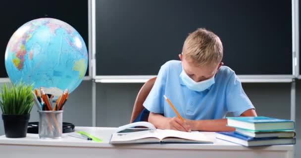 Small Caucasian teen boy in medical mask sitting at desk with world globe in school. Teenage schoolboy doing homework and writing exercise in copybook. Pupil learning in classroom. Coronavirus concept - Footage, Video