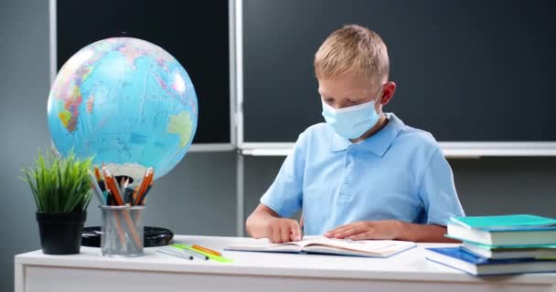 Pieni valkoihoinen teinipoika lääketieteellisessä naamiossa istuu pöydän ääressä maailman kanssa koulussa. Teini-ikäinen koulupoika lukee oppikirjaa ja seuraa sormella. Oppilaat oppivat luokassa. Coronavirus-käsite. - Materiaali, video
