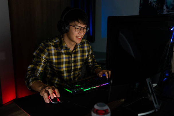Kulaklıklı yakışıklı, heyecanlı Asyalı oyuncu sıcak ve neon ışık, oyun ve teknoloji e-spor konseptiyle donatılmış rahat bir odada bilgisayar oyunu oynarken eğleniyor ve seviniyor.. - Fotoğraf, Görsel