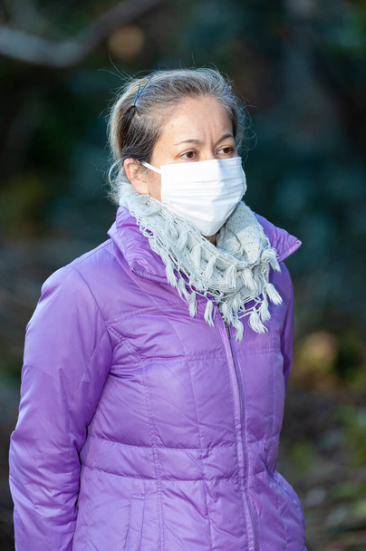 Вид сбоку красивой смешанной расы женщины средних лет в белой маске для защиты от коронавируса (COVID-19) и других инфекционных заболеваний. Размытое прошлое. Копирование пространства. - Фото, изображение