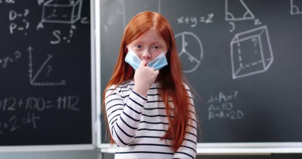 Portret van een schattig, klein blank roodharig meisje met rugzak in de klas, kijkend naar de camera en een medisch masker afnemend. Klein schoolmeisje met rood haar glimlachend op school Coronavirus - Video