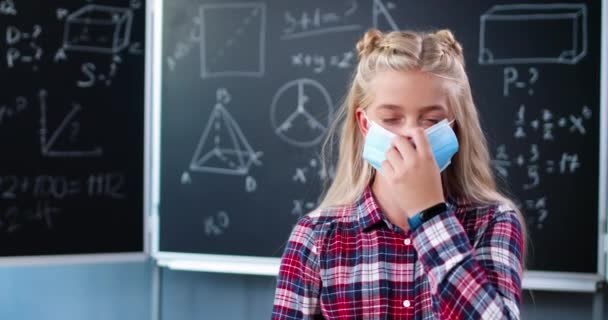 Portrait of pretty happy teen Caucasian girl with fair hair in classroom looking at camera and taking off medical mask. Schoolgirl smiling at blackboard in school. Covid-19 pandemic study concept. - Footage, Video
