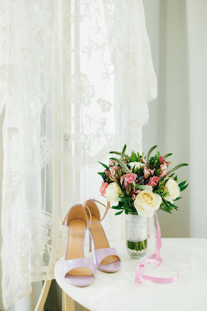 Sandały liliowe na białym stole przy oknie z białym welonem ślubnym i bukietem kwiatów w wazonie w pokoju. - Zdjęcie, obraz