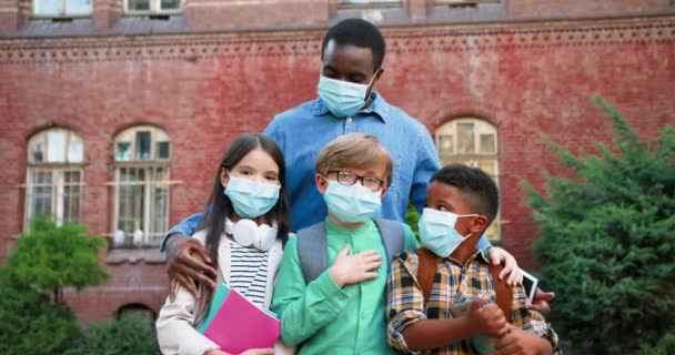 Portrait of mixed-races junior students standing with male teacher near school in masks in lockdown. Cute Caucasian girl and boy. African American boy pupil with copybook outdoor. School concept - Footage, Video
