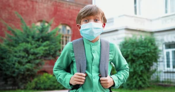 Κοντινό πορτραίτο ενός χαριτωμένου καυκάσιου αγοριού με ιατρική μάσκα μπροστά από το σχολείο. Μαθητής με σχολική τσάντα σε καραντίνα. Μελέτη κατά τη διάρκεια πανδημίας. Έννοια ιού - Πλάνα, βίντεο