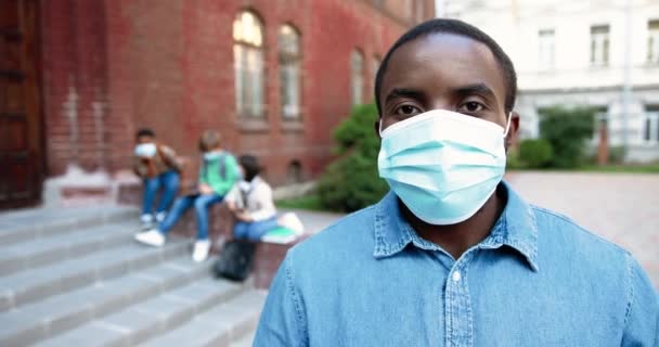 Close up portrait of African American handsome man in mask looking away and smiling to camera near school. Male teacher outdoor. Mixed-races junior students on background. Quarantine concept - Footage, Video