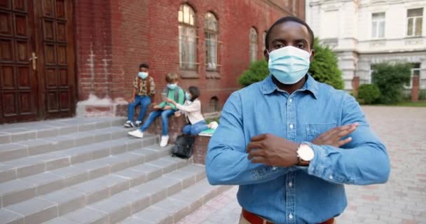 Portrait of African American handsome male teacher in mask looking at camera in front of school. Man standing outdoor at schoolyard. Mixed-races junior students on background. Pandemic concept - Footage, Video
