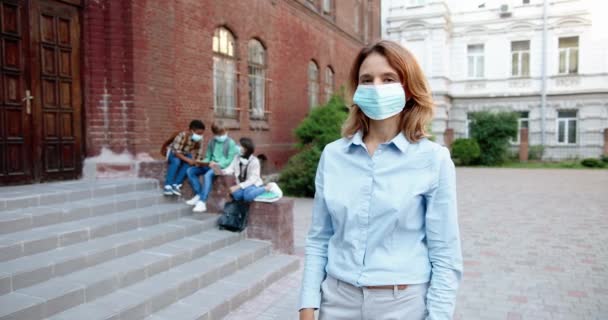 Portrait of beautiful Caucasian happy female teacher in mask smiling to camera in front of school. Woman standing outdoor at schoolyard. Mixed-races junior students on background. Pandemic concept - Footage, Video