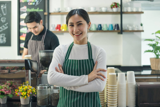 atrakcyjna młoda Azjatka uśmiechnięta i pewna siebie w kawiarni. Młoda baristka prowadzi kawiarnię z rękami złożonymi w przednim barze. Kelnerki i mężczyźni noszą fartuchy pracujące w restauracjach. - Zdjęcie, obraz