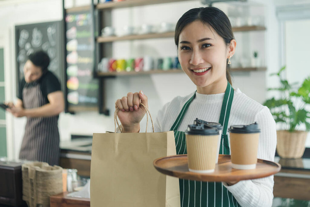 Attraktive junge Asiatin, die im Café selbstbewusst lächelt. junge Barista, Inhaberin eines Cafés, hält Kaffeetasse und Papiertüte vor der Theke. Kellnerin trägt Schürze in Restaurants - Foto, Bild