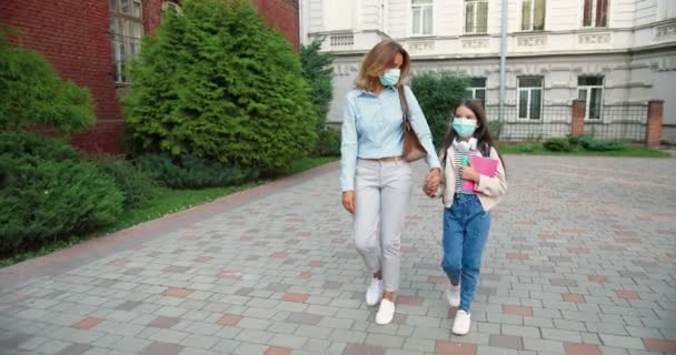 Kaukázusi gyönyörű anya maszkban elviszi a lányát az iskolába a karantén után. Egy aranyos lány fülhallgatóval és másolatokkal az iskolaudvaron. Vissza a suliba. Biztonsági intézkedések. A tudás fogalma - Felvétel, videó