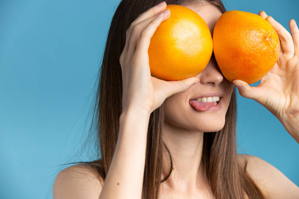 Young awesome girl holding juicy halves of grapefruit against her eyes , sticking out tongue. Blue cut out background - Fotó, kép