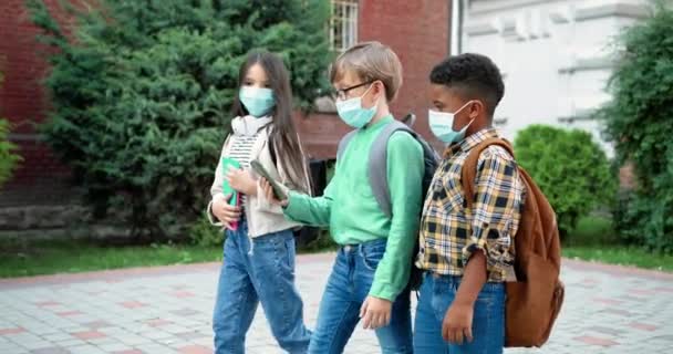 Mnohonárodnostní žáci v maskách s batohy chodí do školy. Běloška s copybookem. Afroameričan se školní brašnou. Dívej se na něco na smartphonu. Studijní koncept - Záběry, video