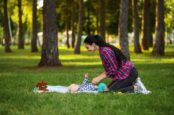 Madre juega con su hijo pequeño en el césped en el parque de verano. Mamá con niños con distracciones en el césped, picnic con niños a cuadros en el bosque, familia de vacaciones - Foto, imagen