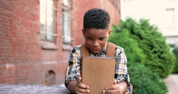 Portré koncentrált afro-amerikai tini fiú kopog és böngészés tabletta szabadban. Aranyos fiú junior diák használ eszközt az iskola közelében, és nézi a képernyőt. Iskolásgyerek használja modul Pupil koncepció - Felvétel, videó
