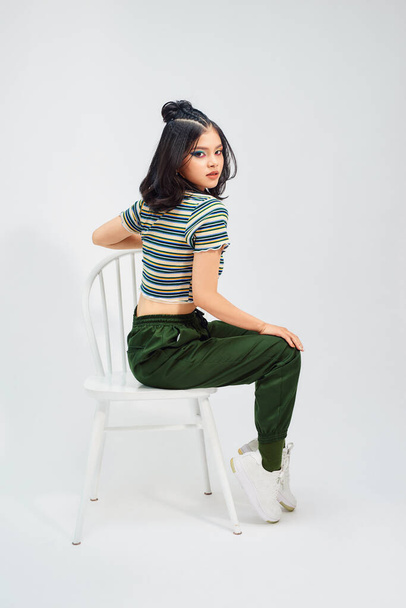 krásná asijská dívka v módní v croptop sedí na stoličce a dívá se na kameru - Fotografie, Obrázek