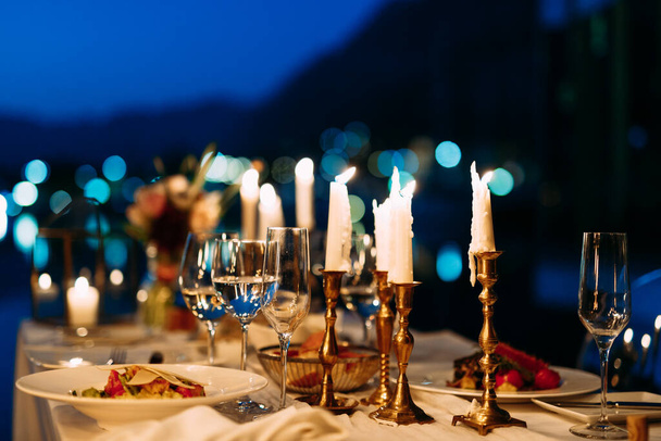 Κεριά σε κηροπήγια με ποτήρια και κεράσματα στο τραπέζι το βράδυ με ρηχό βάθος αγρού. - Φωτογραφία, εικόνα