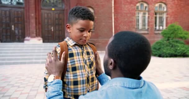 Primer plano retrato de adolescente alumno masculino con mochila hablando con el padre cerca de la escuela antes de las clases. Hombre afroamericano guapo animando a su hijo en el patio de la escuela.Estudiante junior. Concepto exterior - Imágenes, Vídeo
