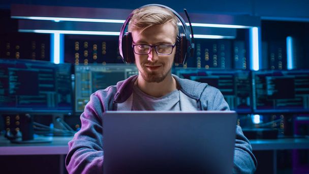 Portrait of Software Developer Hacker Gamer Wearing Glasses and Headset Sitting at His Desk and Working Playing on Laptop. In the Background Dark High Tech Environment with Multiple Displays. - Photo, Image