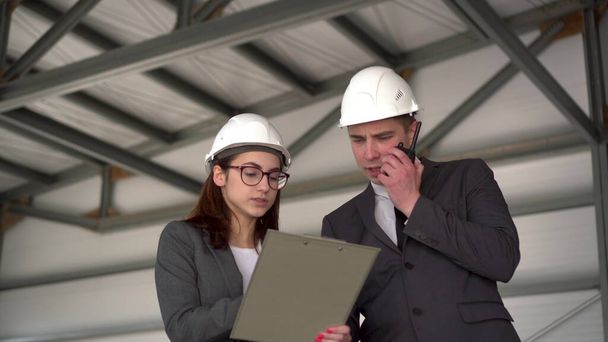Young man and woman in helmets with documents at a construction site. Bosses in suits are discussing an architecture project. A man speaks on a walkie-talkie. People look around. - Photo, Image