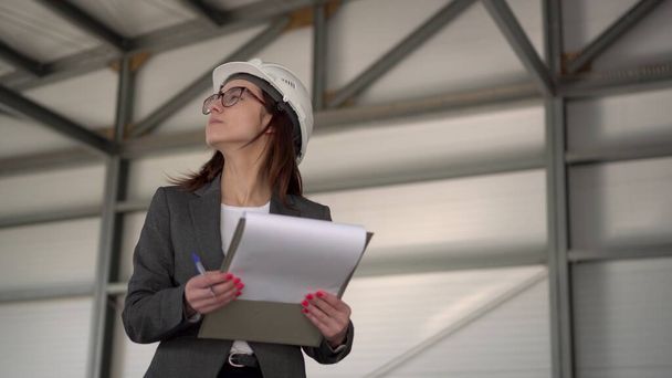 Young woman in a helmet with documents at a construction site. The boss woman in a suit keeps records of documents on architecture. The girl looks around. - Photo, Image