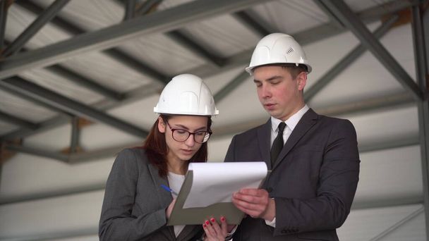 Young man and woman in helmets with documents at a construction site. Businessmen in suits conclude an agreement. A woman signed documents. - Photo, Image