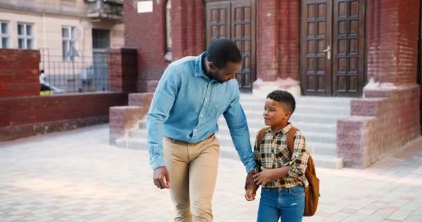 Yakışıklı Afro-Amerikan bir adam okulun yanında yakışıklı bir çocukla yürüyor ve konuşuyor. Sırt çantalı genç bir öğrenci babasına bir şey söylüyor. Baba oğlunu dinliyor. Öğrenci konsepti - Video, Çekim