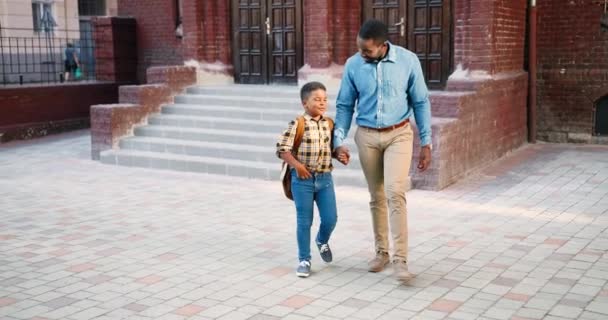 Feliz hombre afroamericano guapo caminando con un lindo colegial cerca de la escuela y hablando. Adolescente estudiante junior con mochila charlando con su padre al aire libre después de las clases. Concepto de conversación - Imágenes, Vídeo