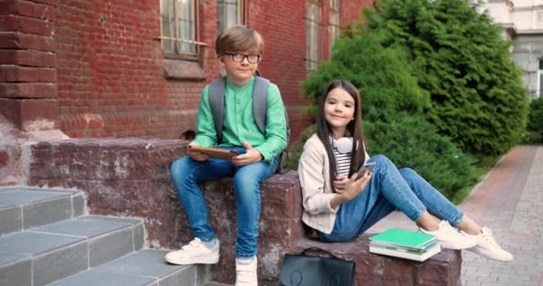Roztomilý kavkazský malý školáček s tabletem a školačka s smartphonem s úsměvem na kameru, zatímco sedí blízko školy venku. Radostní žáci s pomůckami v rukou v dobré náladě na školním dvoře. - Záběry, video