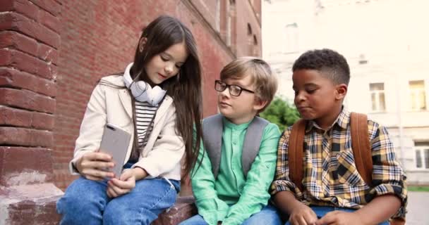 Portret mieszanych dzieci siedzących koło szkoły i rozmawiających na świeżym powietrzu. Biała ślicznotka trzymająca smartfona w ręku i rozmawiająca z chłopakiem w okularach i Afroamerykańskim kolegą z klasy. Koncepcja przyjaciół - Materiał filmowy, wideo