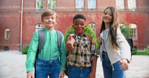 Radostní multietničtí žáci se školními taškami stojící před školou a zvedající palce. Hezká běloška se usmívá venku. Afroameričan roztomilý chlapec v dobré náladě. Koncept portrétu - Záběry, video