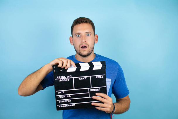 Mladý pohledný muž na sobě modré ležérní tričko přes izolované modré pozadí držení clapperboard strach a překvapení - Fotografie, Obrázek
