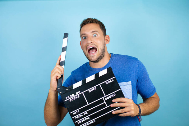 Young handsome man wearing blue casual t-shirt over isolated blue background holding clapperboard very happy - Photo, Image