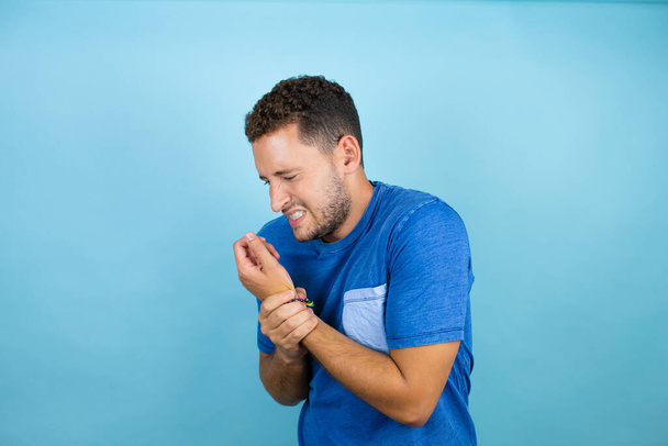 Junger gutaussehender Mann, der ein blaues lässiges T-Shirt über isoliertem blauem Hintergrund trägt, leidet unter Schmerzen an Händen und Fingern, Arthritis-Entzündung - Foto, Bild