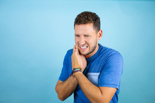 Молодой красивый мужчина в голубой футболке на изолированном синем фоне касается рта рукой с болезненным выражением лица из-за зубной боли или зубной болезни на зубах - Фото, изображение