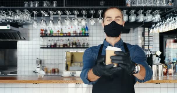 Porträt einer jungen kaukasischen Barkeeperin mit Maske und Handschuhen, die lächelnd eine Tasse Kaffee in die Kamera reicht. Barrista reicht Kaffee to go im Café. Covid-10-Konzept sozialer Distanz. - Filmmaterial, Video