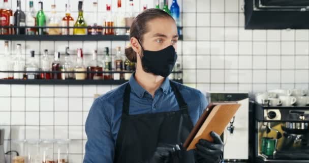 Handsome young Caucasian male waiter in mask, gloves and apron smiling and using tablet device when chatting at bar. Man bartender tapping, scrolling and texting on gadget computer at kitchen. - Footage, Video