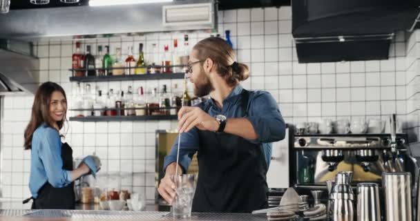 Caucasian young handsome and cheerful man bartender preparing alcohol drink on rocks and smiling to woman colleague. Waiter and waitress talking. Guy barrista making cocktail with ice in bar. - Footage, Video