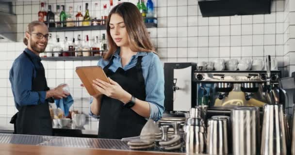Pretty Caucasian female waitress in apron standing in cafe at counter, using tablet device and talking with male waiter. Woman barrista tapping nd scrolling on gadget while chatting with man barman. - Footage, Video