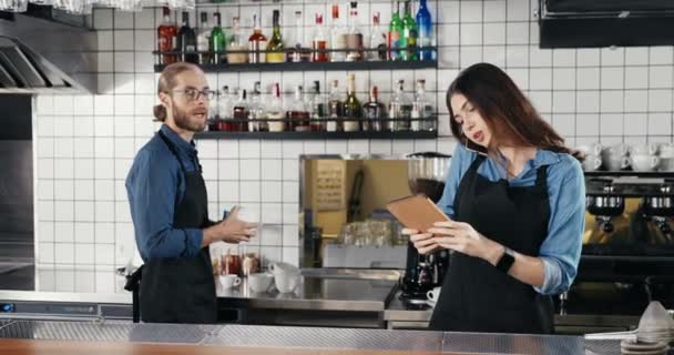 Prachtige jonge vrouwelijke blanke barman die praat op een mobiele telefoon en een tablet vasthoudt aan de bar. Man Barrista werkt op de achtergrond. Serveerster spreekt op mobiele telefoon en tikt op gadget - Video