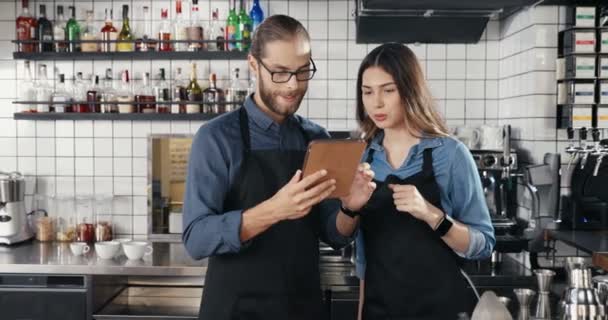 Καυκάσιος νεαρός χαρούμενα αρσενικά και θηλυκά barristas στέκεται στην κουζίνα μπαρ και χρησιμοποιώντας συσκευή tablet. Σερβιτόρος και σερβιτόρα παρακολουθούν βίντεο και πατάνε σε υπολογιστή gadget στο καφέ. Μπάρμαν. - Πλάνα, βίντεο