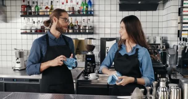 Cheerful young Caucasian couple of male and female barristas in aprons laughing, chatting and wiping glasses in cafe. Happy waiter and waitress cleaning cups and dryin. Barristas working joyfully. - Footage, Video