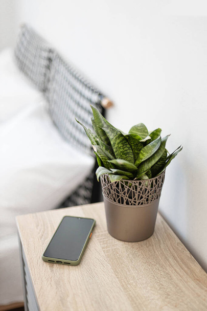 Black smartphone on the nightstand. Green plant in a flowerpot on the table in the bedroom against the background of the bed. Vertical photo orientation - Foto, Imagem