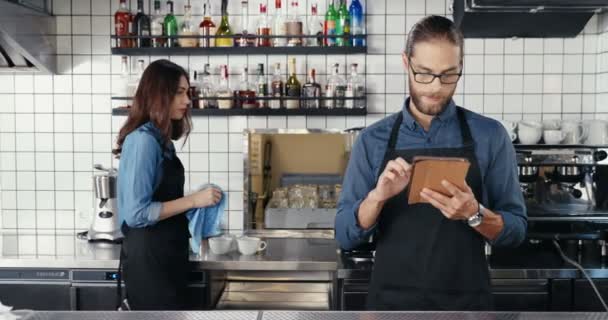 Male Caucasian barmen in glasses and apron using tablet device in cafe. Handsome man waiter in bar tapping and scrolling online on gadget. Waitress working on background. Female wiping glasses. - Footage, Video