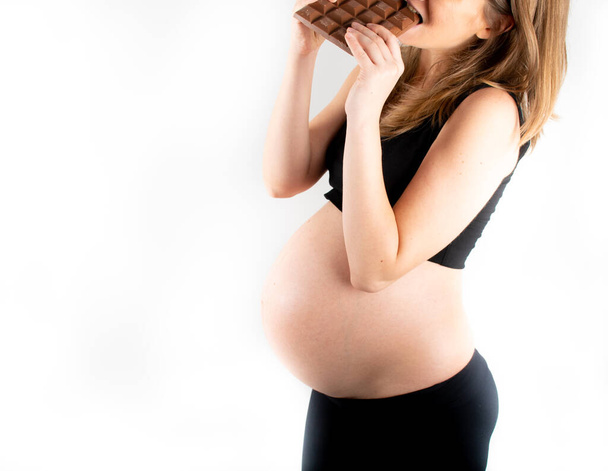 Pregnant woman holding a big dark chocolate bar. Gestational diabetes concept. Sugar free diet, unhealthy, healthy lifestyle. Slim fit skinny woman posing on white background. Black top and leggings - Foto, imagen