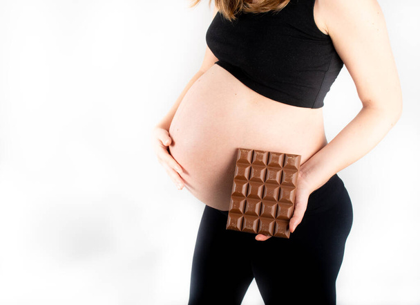 Pregnant woman holding a big dark chocolate bar. Gestational diabetes concept. Sugar free diet, unhealthy, healthy lifestyle. Slim fit skinny woman posing on white background. Black top and leggings - 写真・画像